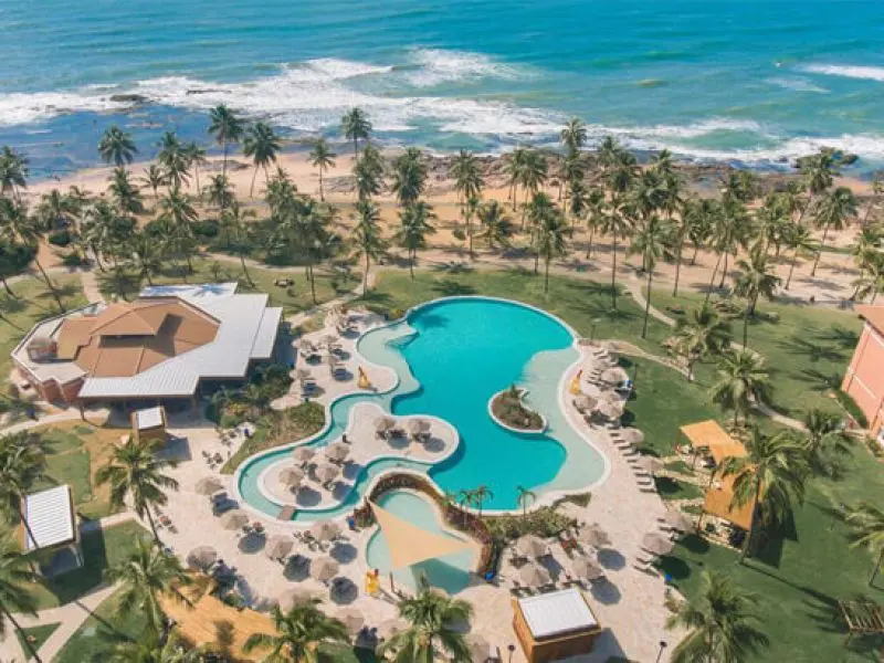 Imagem de um complexo turístico, próximo de uma praia e uma pequena vegetação verde, com piscina e alguns prédios