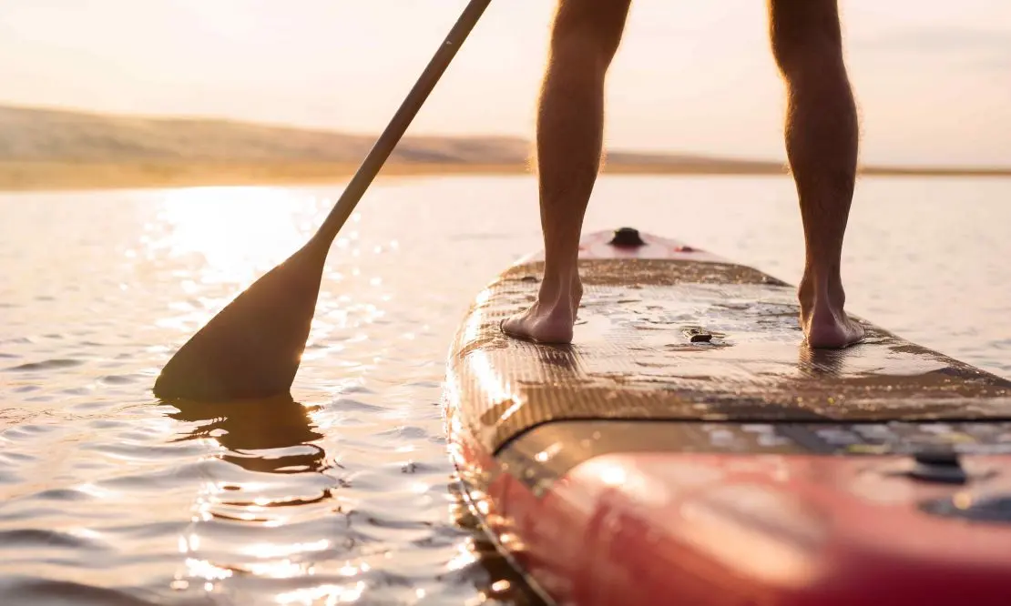 Parte das pernas de uma pessoa em cima de uma prancha de Stand up paddle, remando para o sol que está se pondo