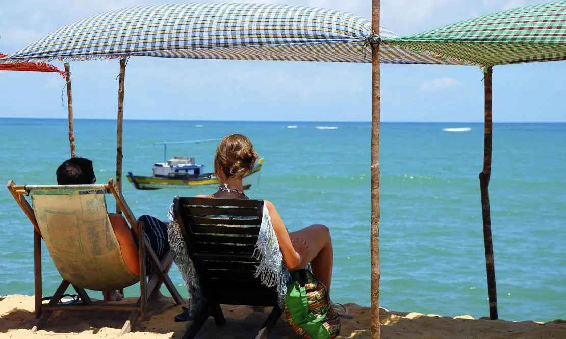 Casal sentado em espreguiçadeiras de madeira, de frente para o mar azul de Trancoso