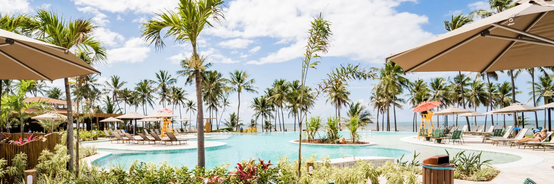 Imagem de uma piscina num complexo turístico próximo a praia e a uma vegetação, com espreguiçadeiras e guarda-sóis 