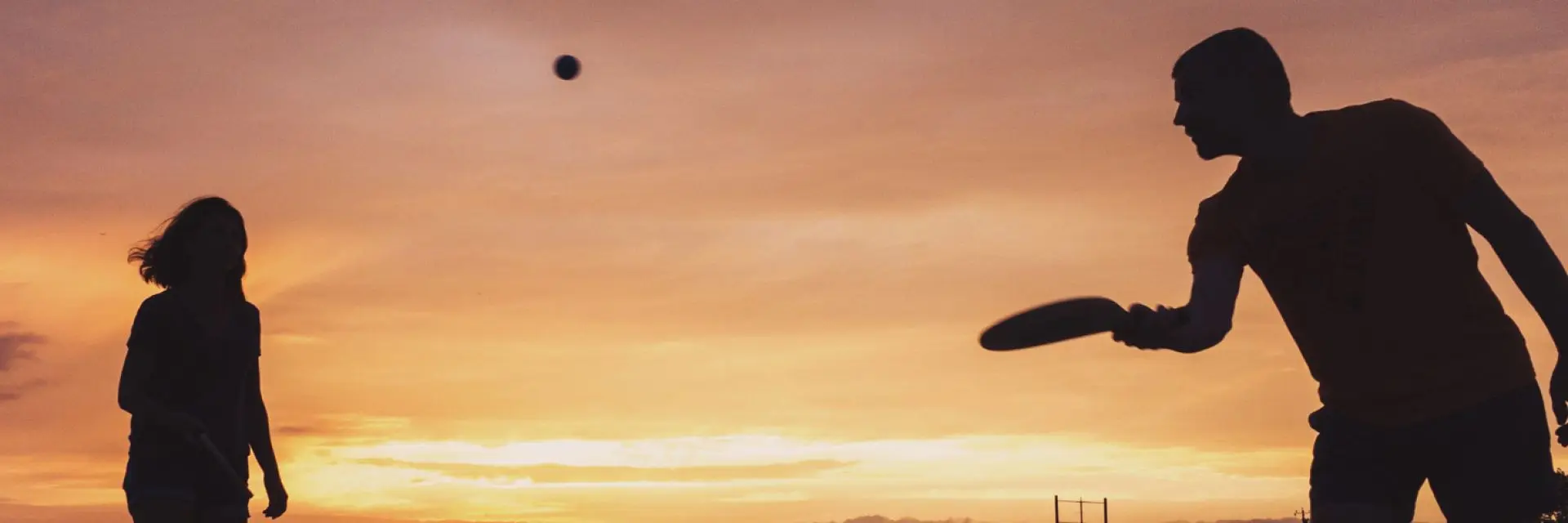 Imagem da sombra de duas pessoas jogando frescobol numa praia com o céu alaranjado e amarelado e o sol se pondo.