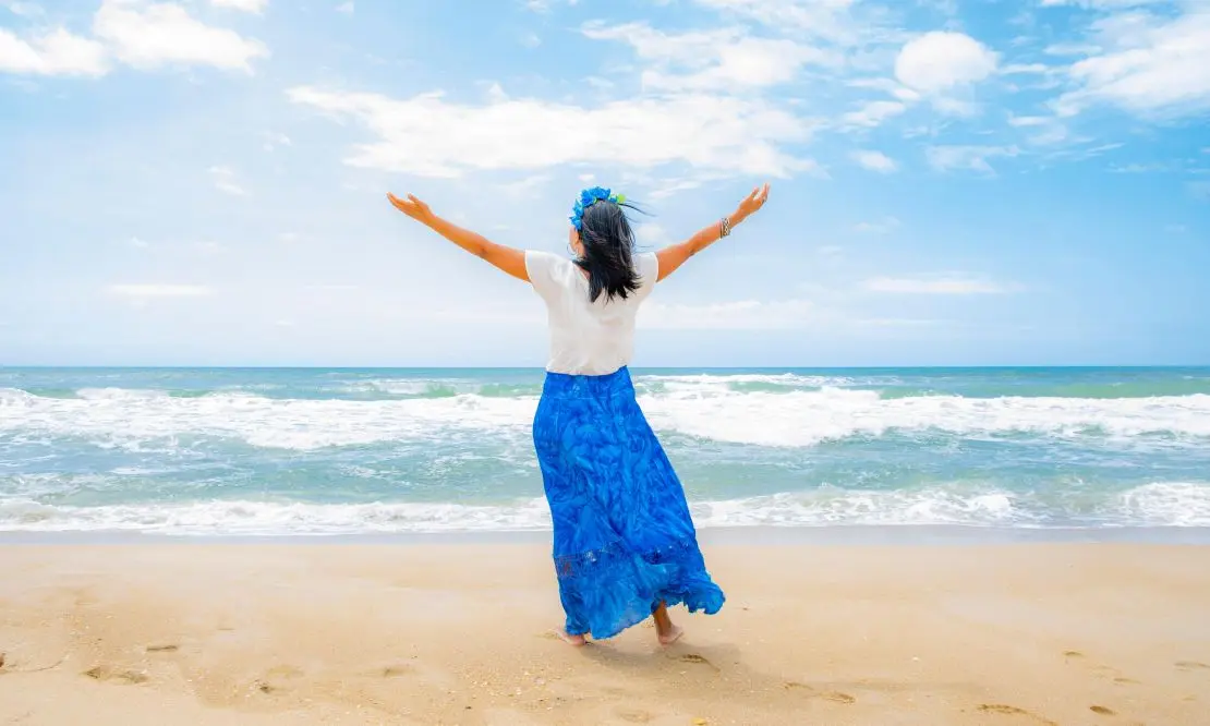 Mulher de costas usando saia longa azul e camiseta branca, olhando para o mar de braçoa abertos